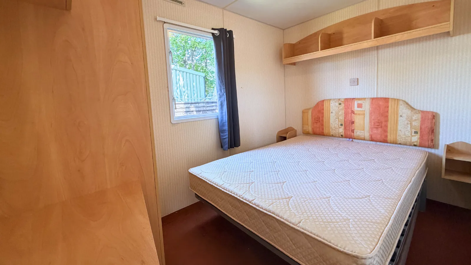 An interior view of a static caravan bedroom with a large bed and overhead shelfing with a window att the side